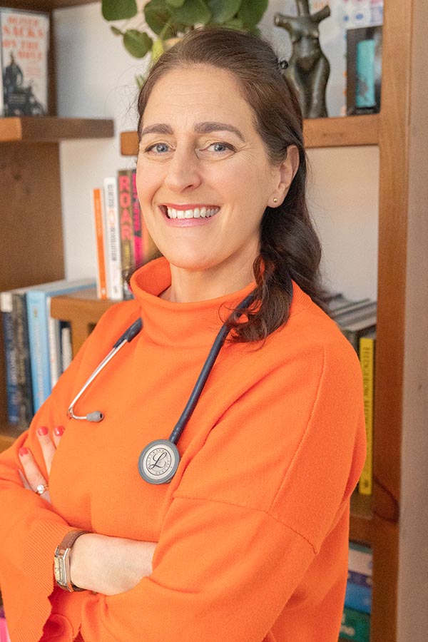 A picture of Alex smiling towards the camera wearing an orange jumper with a stethoscope around her neck.